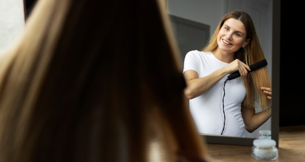 woman-straightening-hair-with-flat-iron-smiling-in-mirror smiling woman in white shirt using flat iron in front of mirror